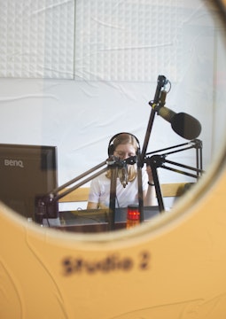 A travers une porte vitrée, une femme dans un studio radio, parle dans le micro avec un casque sur les oreilles