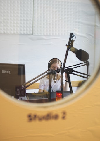 A travers une porte vitrée, une femme dans un studio radio, parle dans le micro avec un casque sur les oreilles